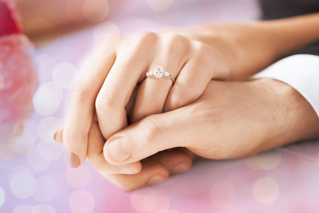 people, holidays, engagement and love concept - close up of engaged couple holding hands with diamond ring over holidays lights background